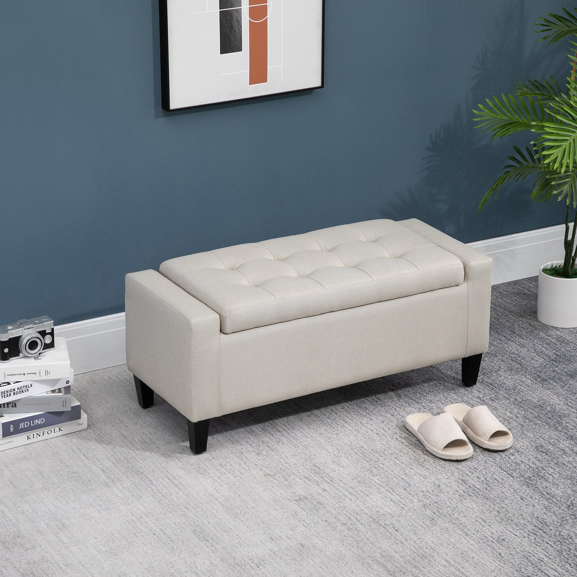 Beige tufted storage bench in a room with a blue wall, books, and a plant.