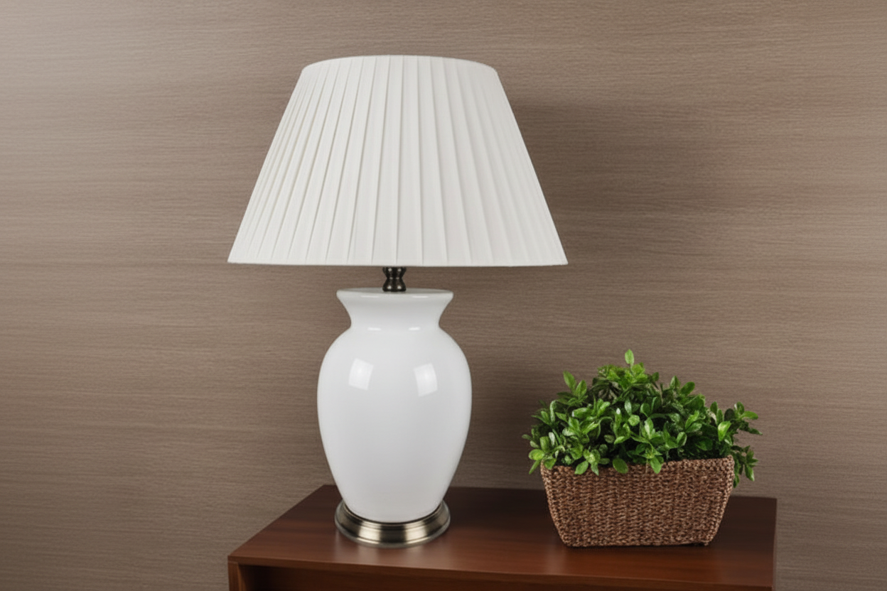 White table lamp with pleated shade on a wooden side table against a brown wall.